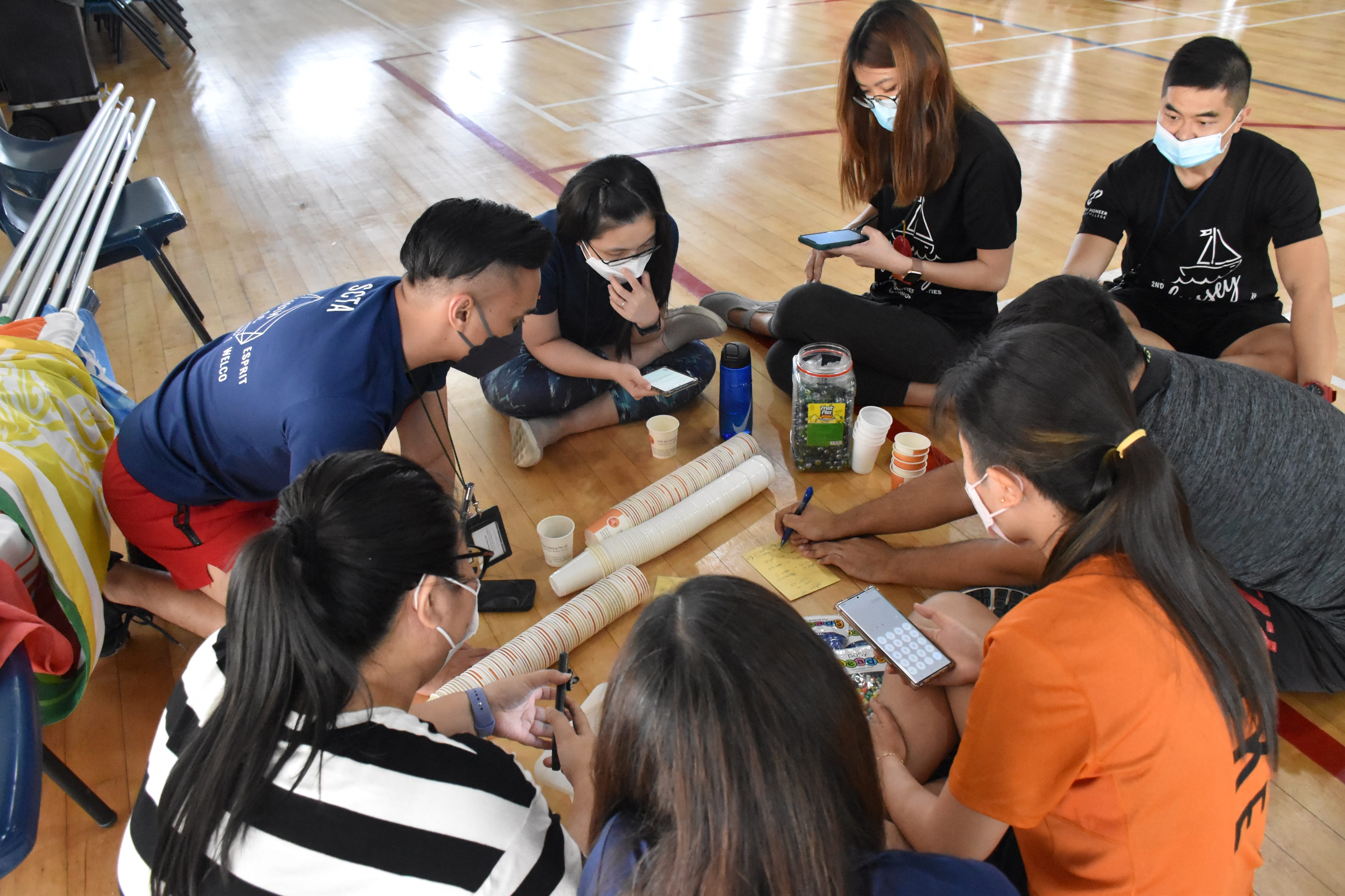 Seven masked people sit in a circle on a gym floor, collaborating with cups, papers, phones, and a jar.